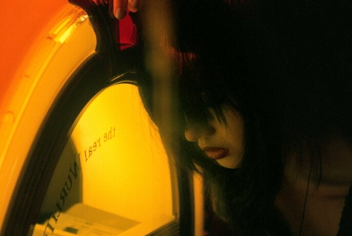 A face partially obscured by hair over the eyes and cheeks, staring into a brightly lit jukebox.