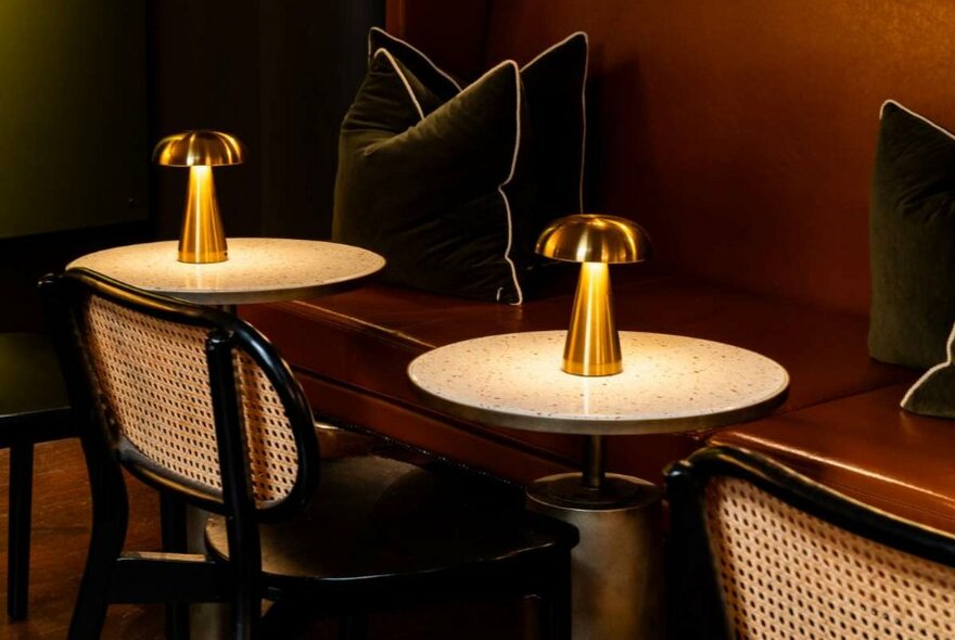 Banquette brown leather seating, small round tables with lamps and rattan dining chairs inside a restaurant.
