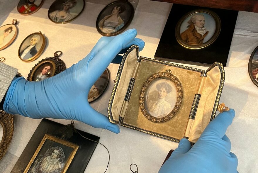 Two hands wearing protective blue gloves holding up a small portrait mounted in a display case. 