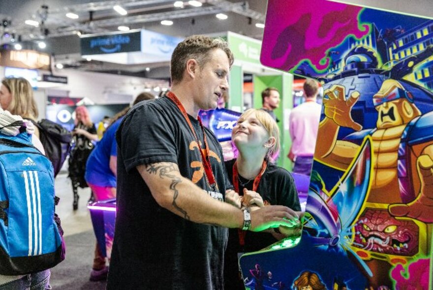 Father and daughter standing at an arcade game console in a crowded exhibition hall.