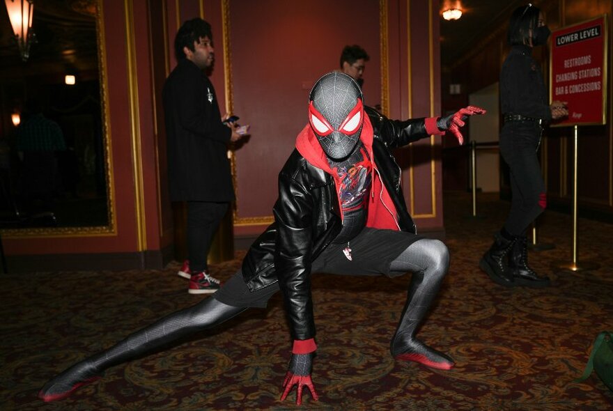 A person dressed as Spiderman in a classic stretched pose in a carpeted foyer with people lining up behind. 