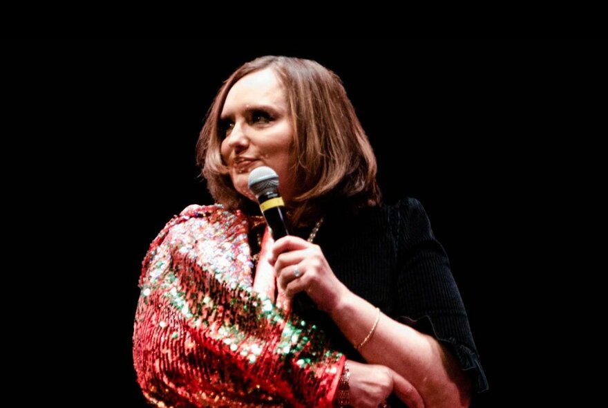 Middle-aged woman holding a microphone up to her mouth, with a sequined shawl draped over her shoulder, on a dark stage.