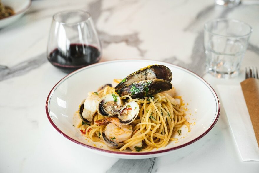 A white dish of spaghetti with clams and mussels, and a glass of red wine, on a table.