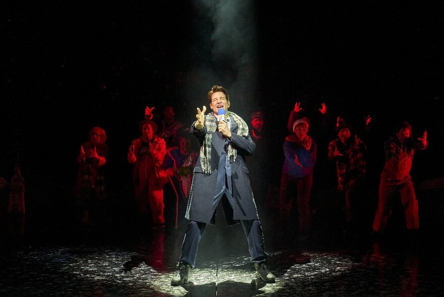 An actor in a suit and coat singing on a darkened theatre stage.
