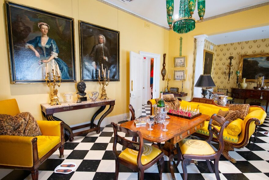 A yellow room inside a historic house with antique furniture there are two paintings of people on the wall.