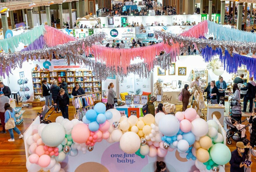 Large expo hall with pink, light blue and silver garlands, balloons in the foreground and people looking at stalls with products on display.