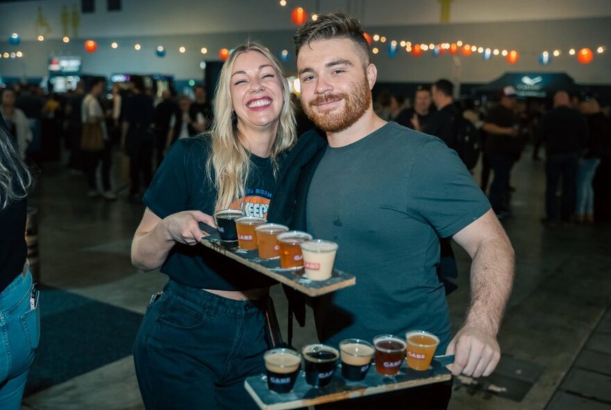 Two people smiling broadly with a beer flight each in a room with coloured fairy lights. 