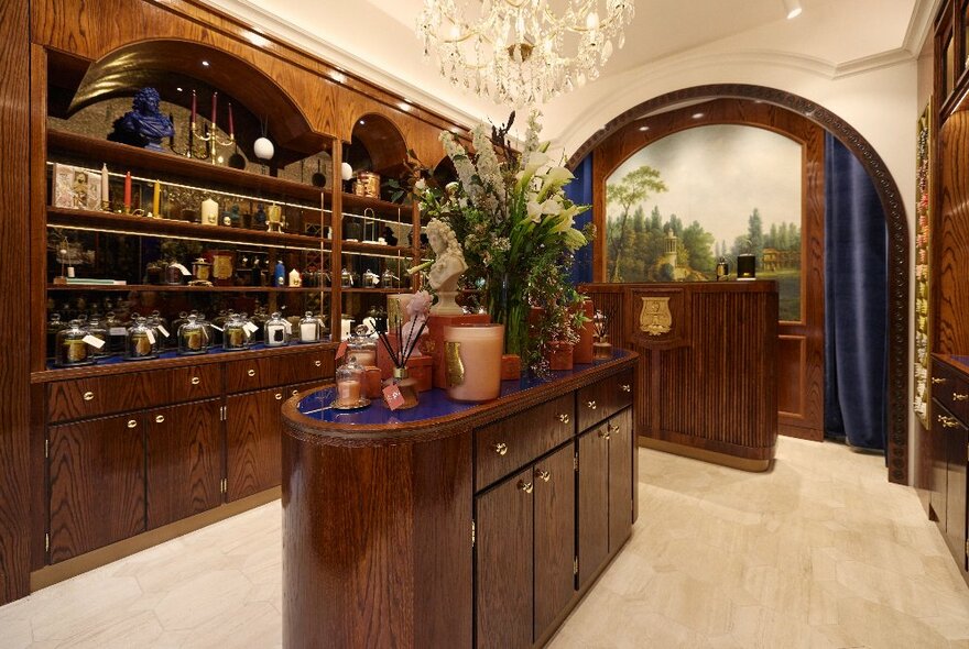 A glamorous candle shop with polished wooden shelving, an island display, a chandelier and blue velvet curtains.