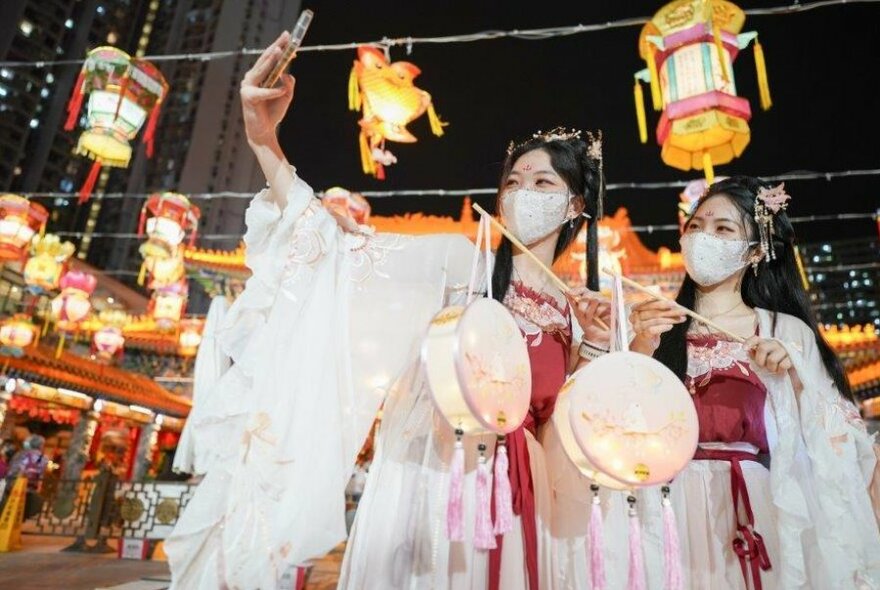 Dancers wearing traditional gowns taking a selfie under hanging lanterns.