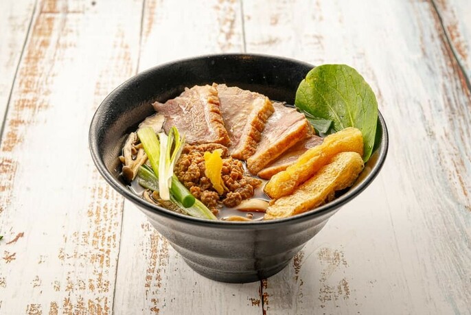 Distressed wooden table with black bowl of ramen topped with pork and garnishes.
