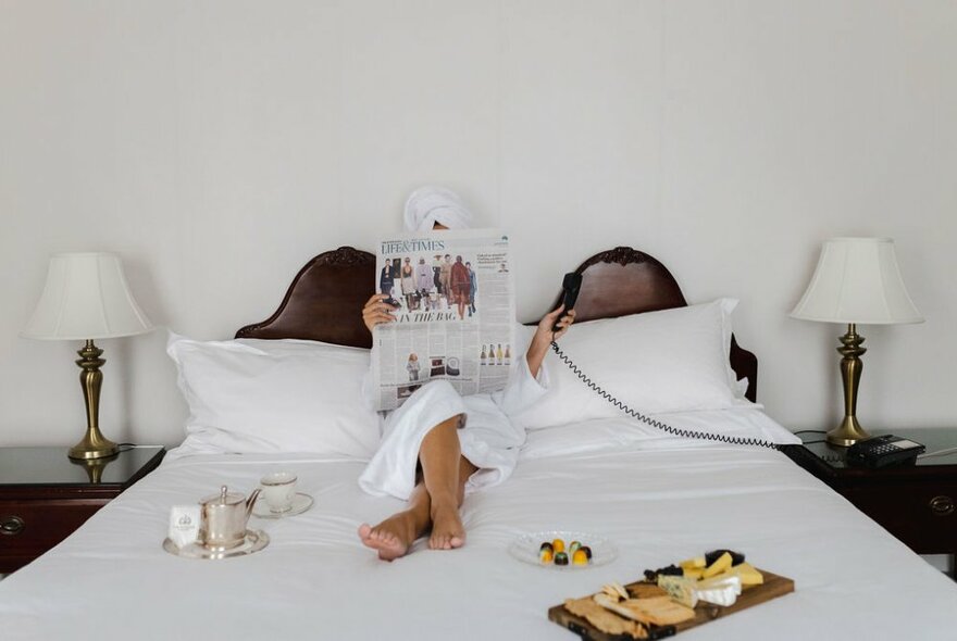 Person reclining back on pillows on a large all white bed in a hotel room, face obscured with a newspaper they hold in their hands, two bedside lamps either side, and a tray of food resting on the edge of the bed.