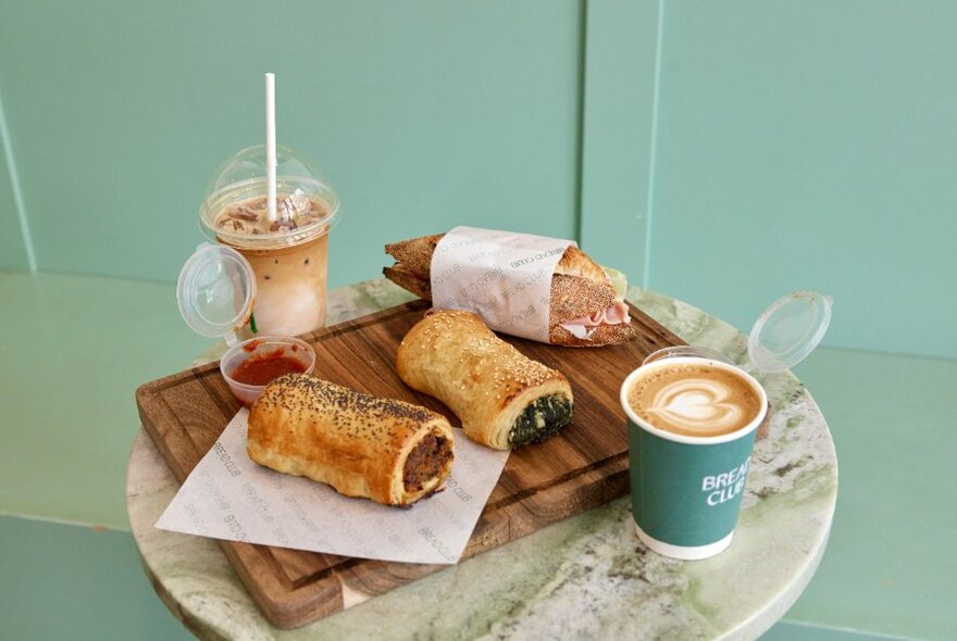 A display of two filled savoury baked pastry rolls, half a filled baguette sandwich, a takeaway coffee and an iced cold drink, resting on a table in a cafe.