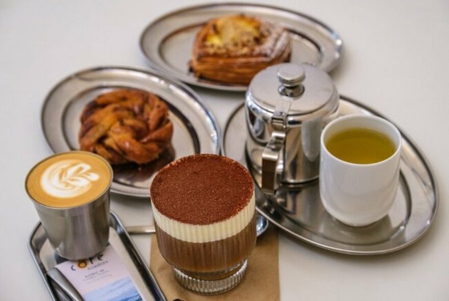A selection of beverages, including a pot of tea, plus two pastries on plates, on a table.