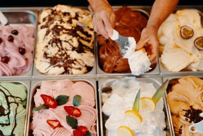 Tubs of different gelato in a glass display cabinet with a pair of hands using a spoon to fill a ice-cream cone with lemon flavored gelato.