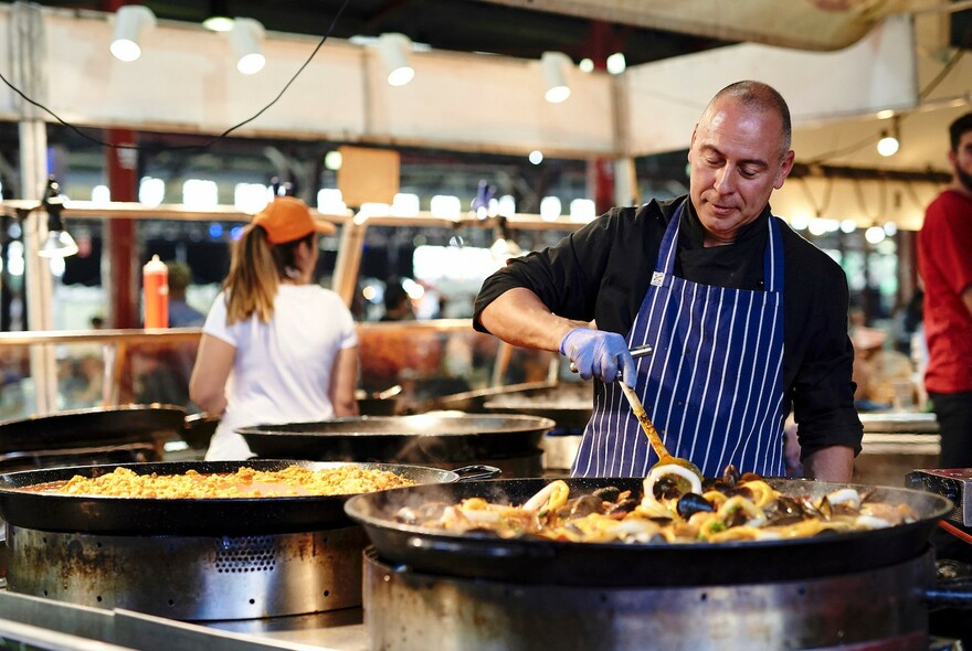 Chef standing over a large group of paella pans, stirring the food. 