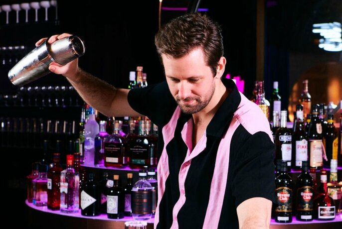 A bartended in a pink and black striped shirt shaking a cocktail 