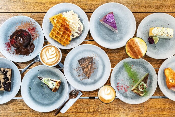 Bird's-eye view of multiple plates of desserts, plus several coffees on a wooden surface.