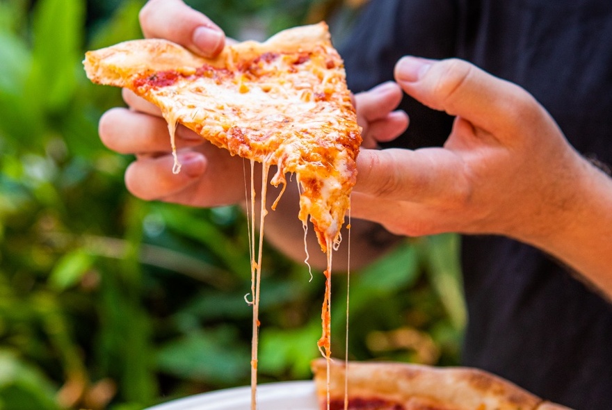 Hand holding up a slice of pizza that is oozing melted cheese.
