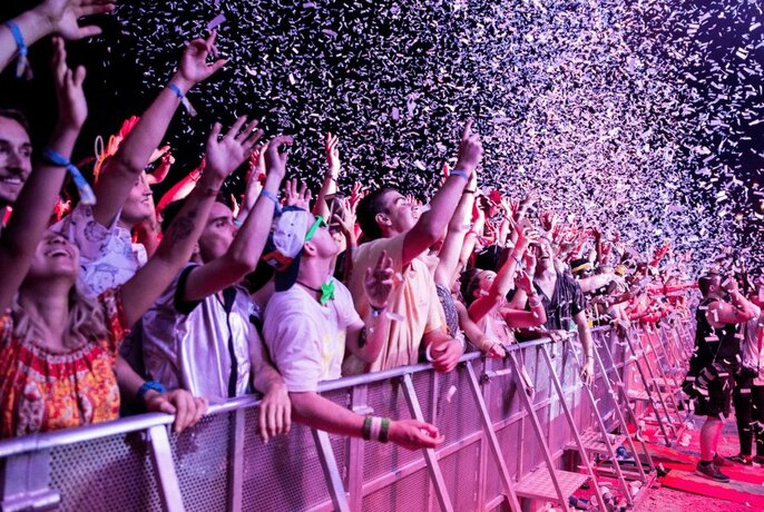 A festival crowd behind barriers, the air full of confetti.