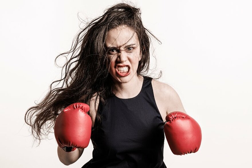 Woman with long, dark hair flowing to side, with gritted teeth, wearing black tank-top and red boxing gloves.