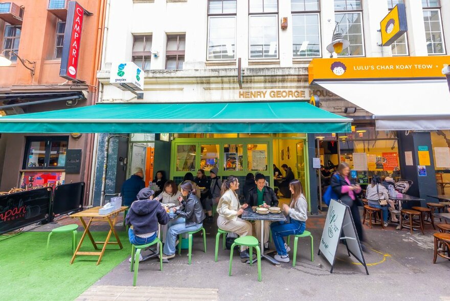 Exterior of Pandan Dessert Bar showing people at an outdoor seating on low stools under a green shade awning, old building in the background.