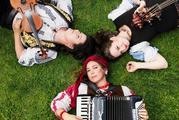 Three musicians of the musical group Vardos lying on grass, holding their instruments of a piano accordion, a violin and a double bass.