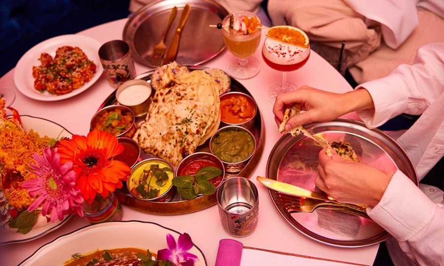 Birds-eye view shot of table full of food at restaurant with two people's hands reaching to dip their naan in dips.
