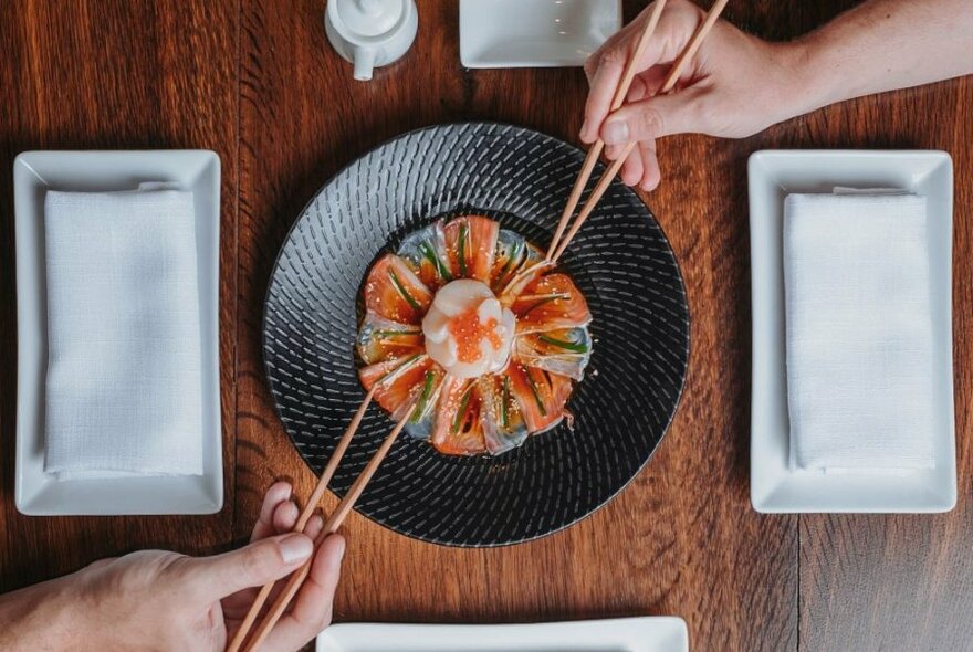 Hands using chopsticks to select salmon sashimi from a dark ceramic plate.