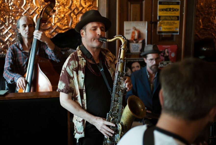 A man in a hat playing saxophone with a man in playing upright bass in the background with people looking on. 