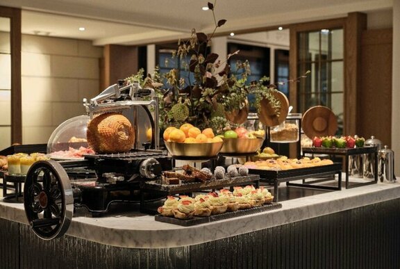 Hotel breakfast bar with ham, pastries, fruit and floral centrepiece.