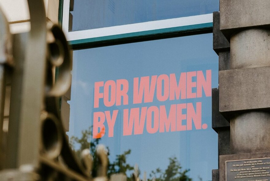 A sign in a window of a heritage building that states in red capital letters, 'FOR WOMEN BY WOMEN'.