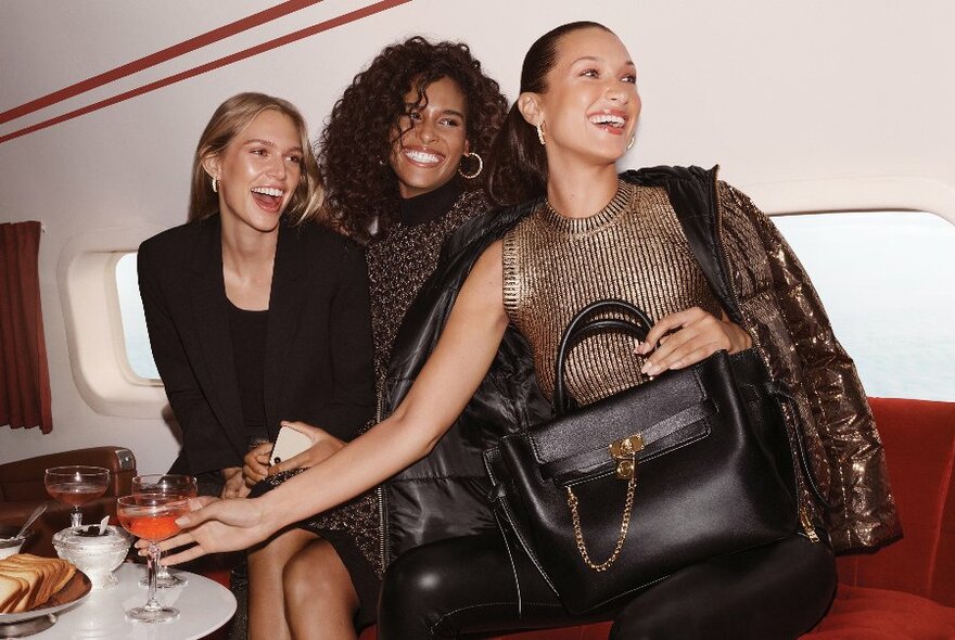 Three models, smiling in black and gold clothes and accessories, seated on red banquette with small windows behind.