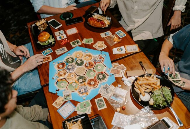 A wooden table with a board game in play on top. People are sitting around the table eating bar snacks and pub food.
