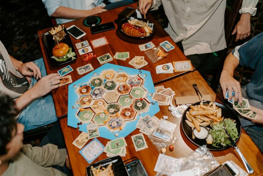 A wooden table with a board game in play on top. People are sitting around the table eating bar snacks and pub food.