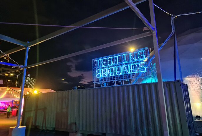 Blue neon signage that spells out Testing Grounds above a shipping container, with a night sky visible and a lit up marquee in the background.