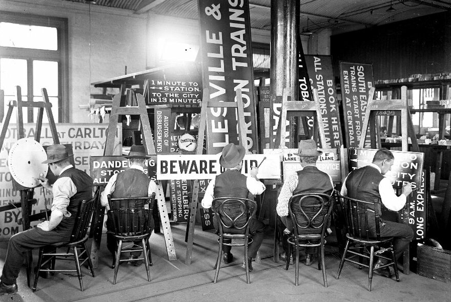 Black and white image of backs of five sign writers at work, facing signs of different sizes. 
