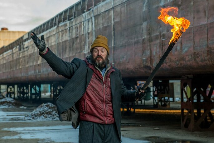 A man outdoors in an industrial setting holding up a fire stick with his arms raised in the air.