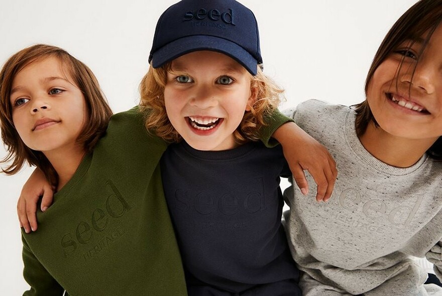 Three children arm in arm wearing casual Seed-logo sweatshirts and a cap.