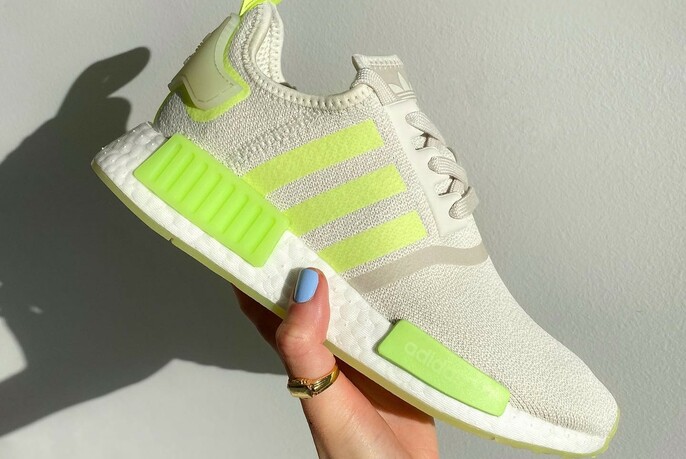 A hand with pale blue nail polish holding an Adidas sneaker with fluorescent lime stripes and details.