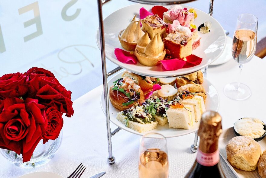 Two-tiers of a tiered serving tray with a selection of treats, alongside a small vase of red roses and champagne.