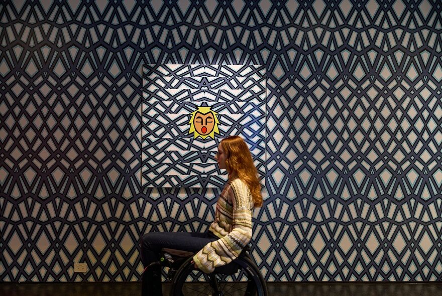 A woman moving past a full-wall artwork of blue geometric pattern with a yellow face in the middle, she is using a wheelchair.