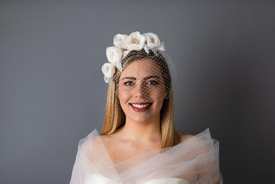 Millinery model standing in front of a grey background, wearing a white floral net fascinator and white tulle gown.