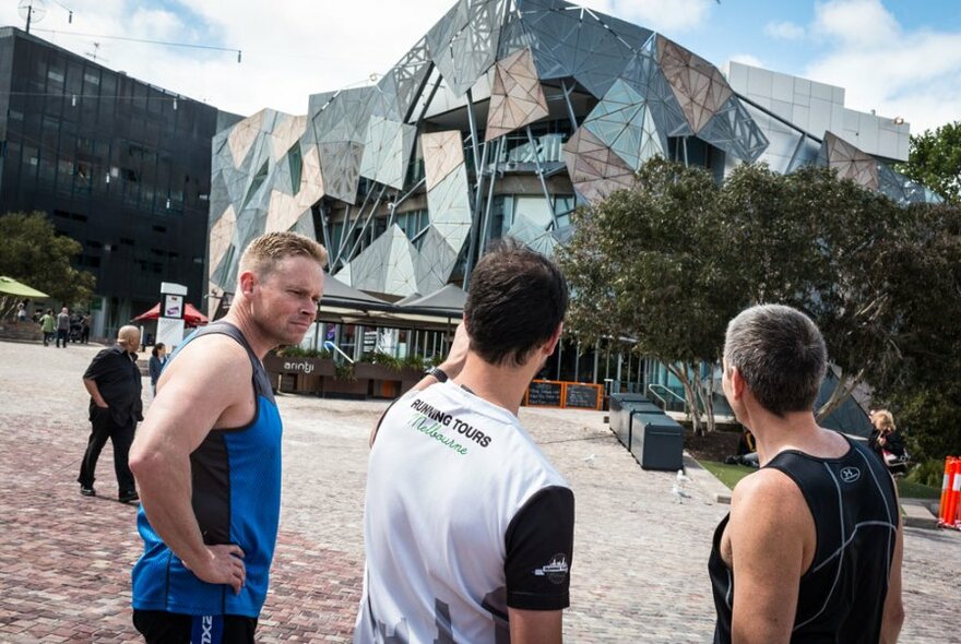 Three people in running active wear standing and looking at a building in Federation Square.