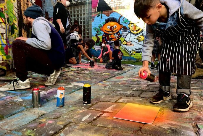 Young person bending over and spraying red and orange canvas on bricks, with others spraypainting to left. 