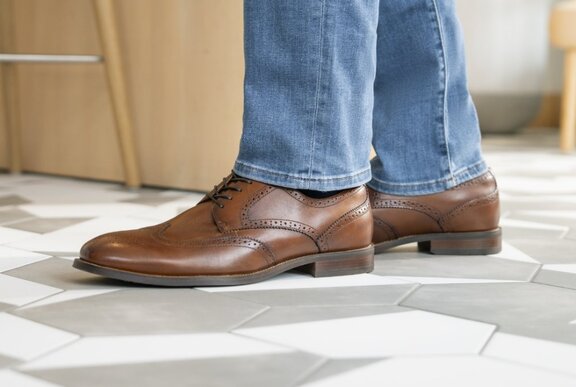 Person standing on tiled floor wearing light wash blue jeans and brown leather shoes.