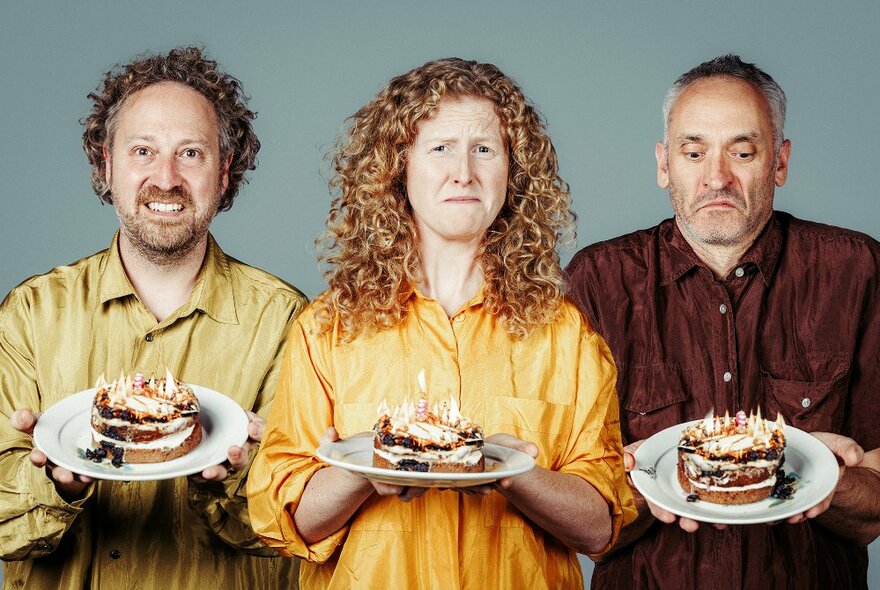 Three performers all facing the camera looking slightly nervous, and each holding a cake with burning candles on it, on a plate in front of them.