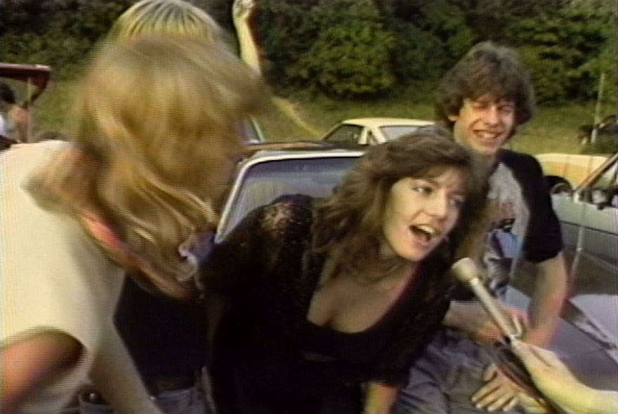 A still from a 1980s video of three young adults, sitting on a car outdoors, one of them leaning forward to talk into a microphone.