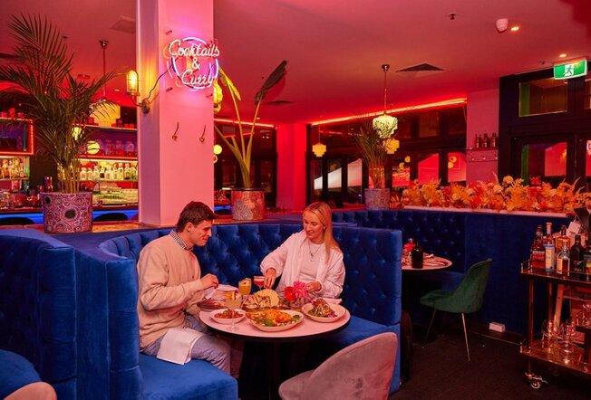 A couple dining on shared Indian dishes in a blue velvet booth in a pink restaurant.