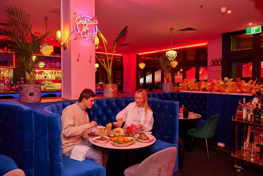 A couple dining on shared Indian dishes in a blue velvet booth in a pink restaurant.