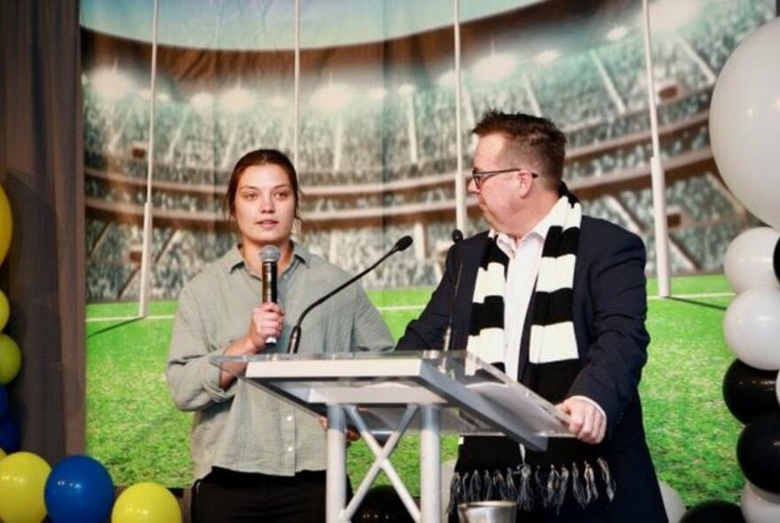 Two people on a stage, standing at a lectern, one of them speaking into a microphone, balloons at the side, and a picture of a the MCG sports stadium behind them on a screen.